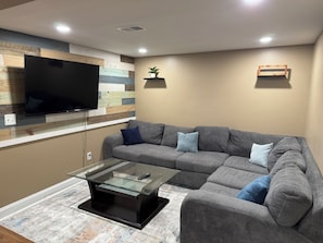 Ein modernes Wohnzimmer mit einem Ecksofa, einem flachbildfernseher, der an einer holzverkleideten Wand montiert ist, einem Glastisch und einem kleinen Topfpflanzen auf einem Regal.