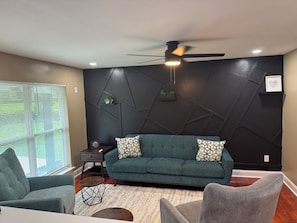 Ein Wohnzimmer mit dunkler Wand, einem grünen Sofa, einem Deckenventilator und einem Fenster mit Jalousien.