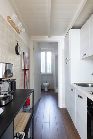 Een smalle keuken met een zwart aanrecht, een houten snijplank, een spoelbak en een raam dat uitkomt op een badkamer.