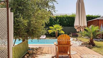 A wooden deck with a chair and a table, a pool, and a green fence.