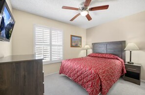 A bedroom with a bed, a ceiling fan, a dresser, a nightstand with a lamp, and a window with blinds.