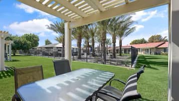 A covered patio with a table and chairs, palm trees, and a fenced area in the background.