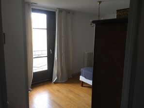 A bedroom with a wooden floor, a radiator, a bed with a blue cover, and a window with curtains. - On the banks of the canal surrounding the town center, very bright apartment (Pamiers)