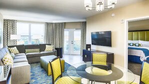 Habitación de hotel con sofá, mesa de comedor con sillas, televisor y balcón con cortinas.