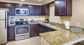 Eine KĂŒche mit dunklen HolzschrĂ€nken, einem EdelstahlkĂŒhlschrank, einem eingebauten Backofen, einer Mikrowelle, Granitarbeitsplatten und einem SpĂŒlbecken.