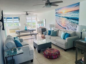Ein Wohnzimmer mit Sofa, Couchtisch und Hocker, ein Essbereich mit Tisch und Stühlen sowie ein großes Gemälde mit Strandmotiv.