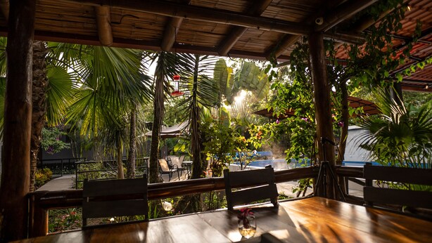 Ein Holztisch mit einer Bank, einer hängenden Leuchte und Blick auf einen Garten mit Palmen und einem blauen Bauwerk.