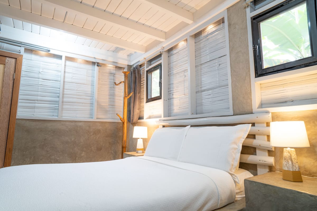 A bedroom with a bed, two bedside tables with lamps, a window with blinds, and a wooden headboard.