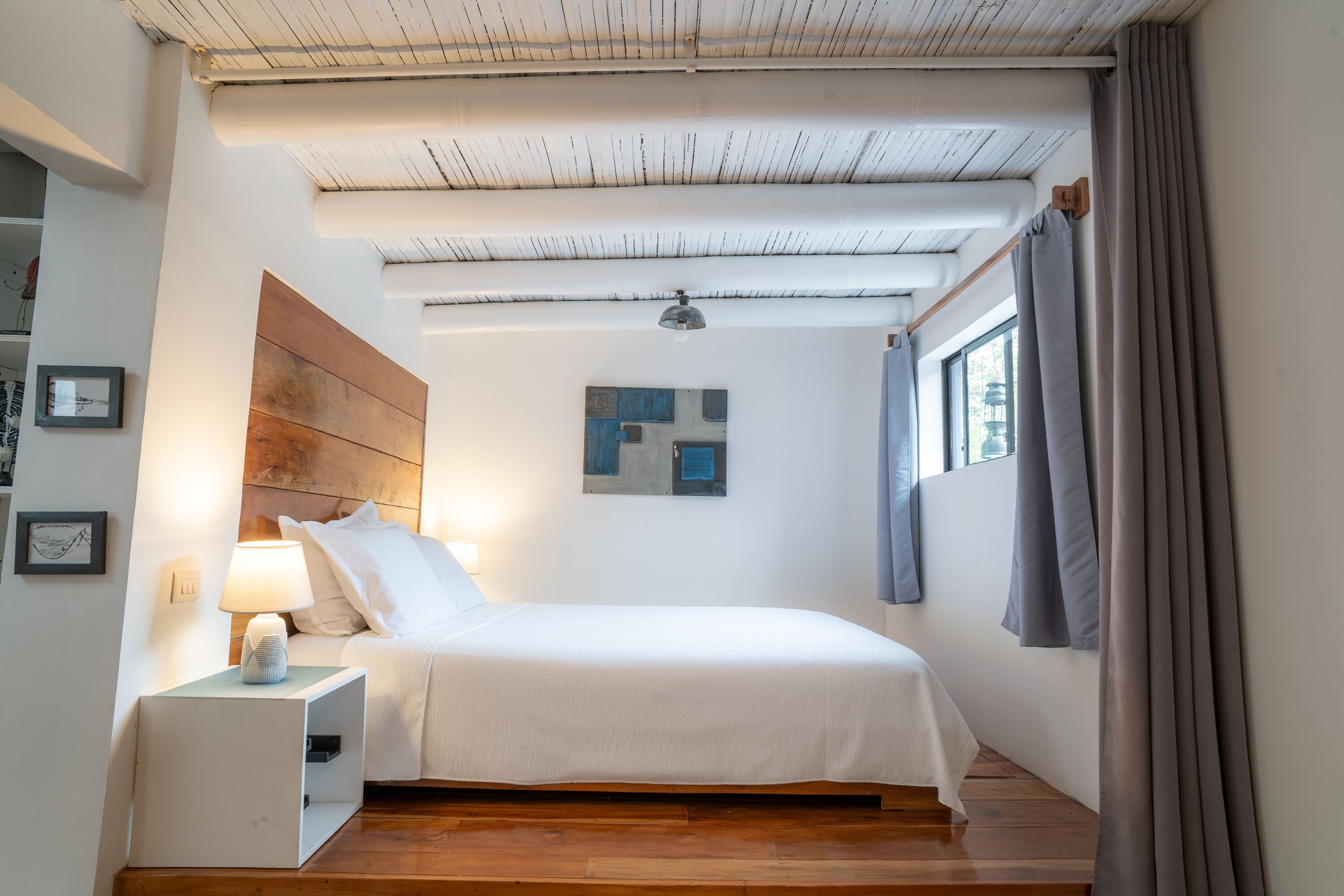 A bedroom with a bed, bedside table, lamp, artwork, and a window with curtains.