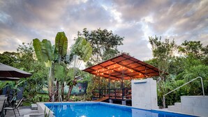 Ein Pool mit Holzterrasse, umgeben von üppiger Vegetation und einem überdachten Sitzbereich.