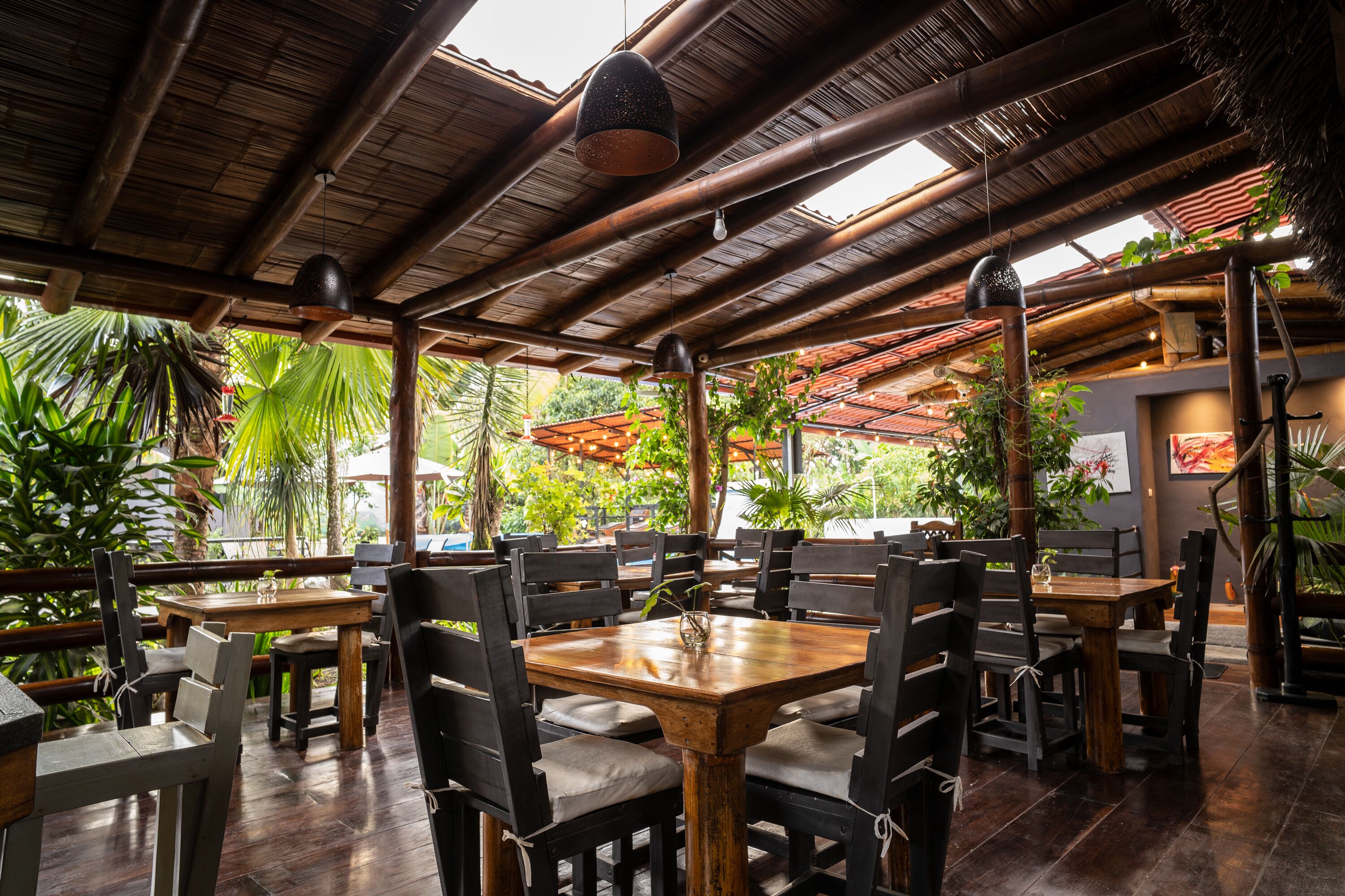 An outdoor restaurant with wooden tables and chairs, bamboo ceiling beams, and tropical plants.
