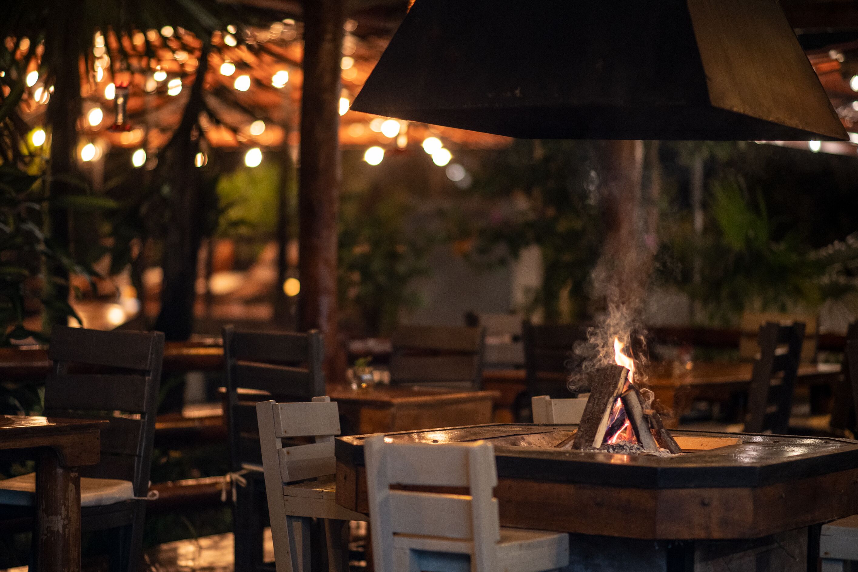 A restaurant with wooden tables and chairs, a central fire feature, and string lights in the background.