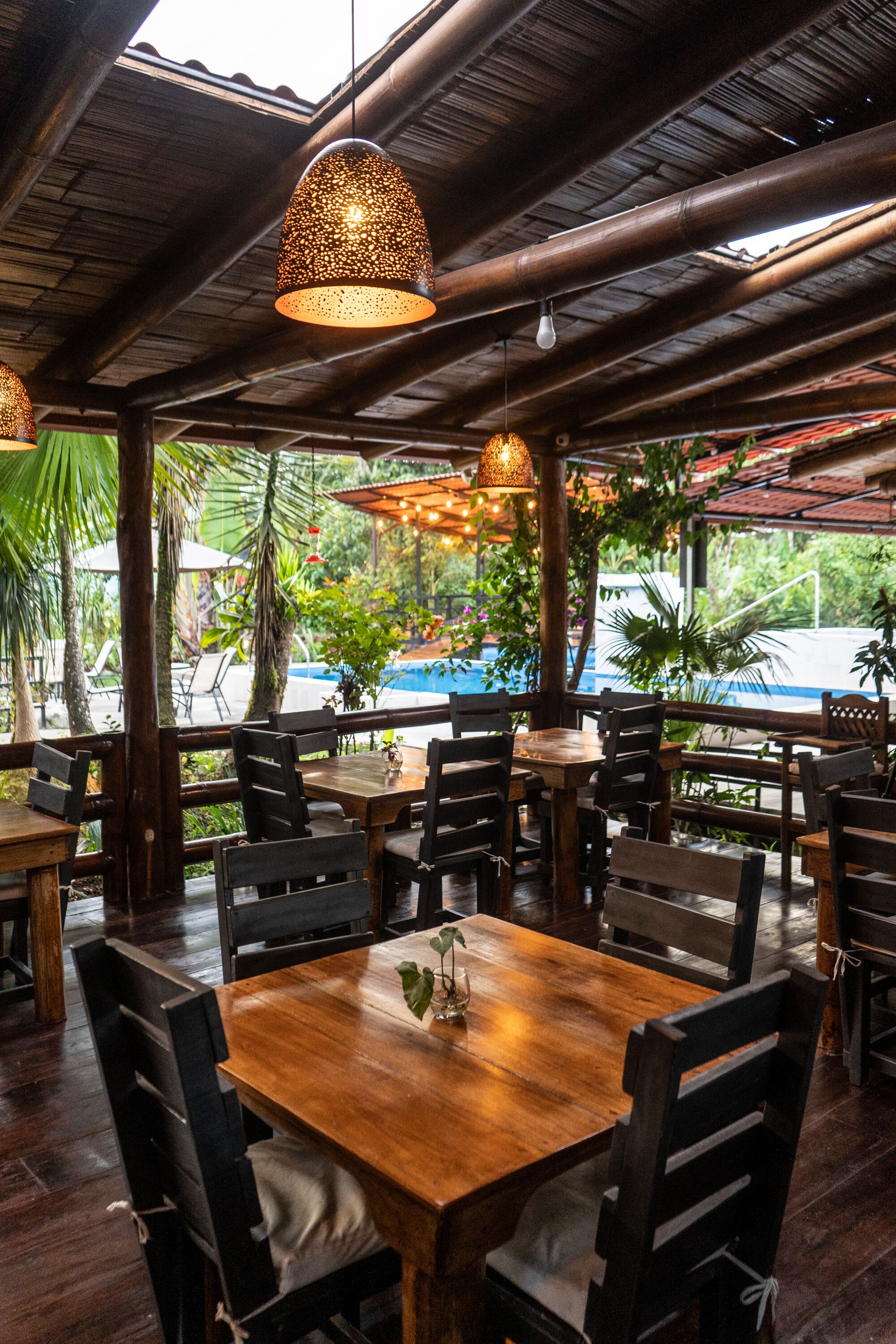 An outdoor restaurant with wooden tables and chairs, hanging lights, and a view of a pool.