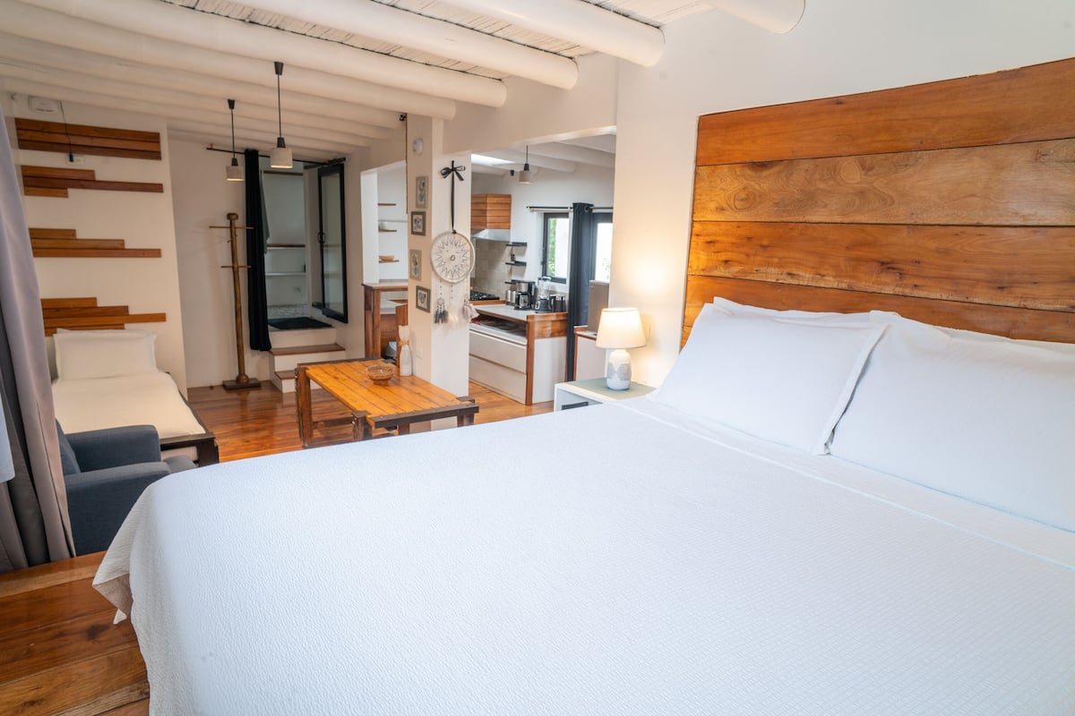A modern hotel room with a large bed, a wooden headboard, a small table, and a view into the kitchen area.