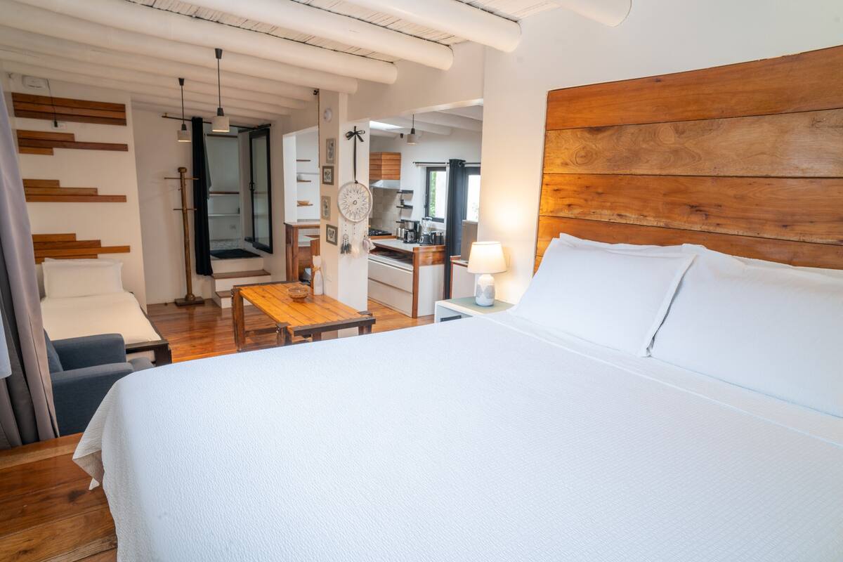 A modern hotel room with a large bed, a wooden headboard, a small table, and a view into the kitchen area.