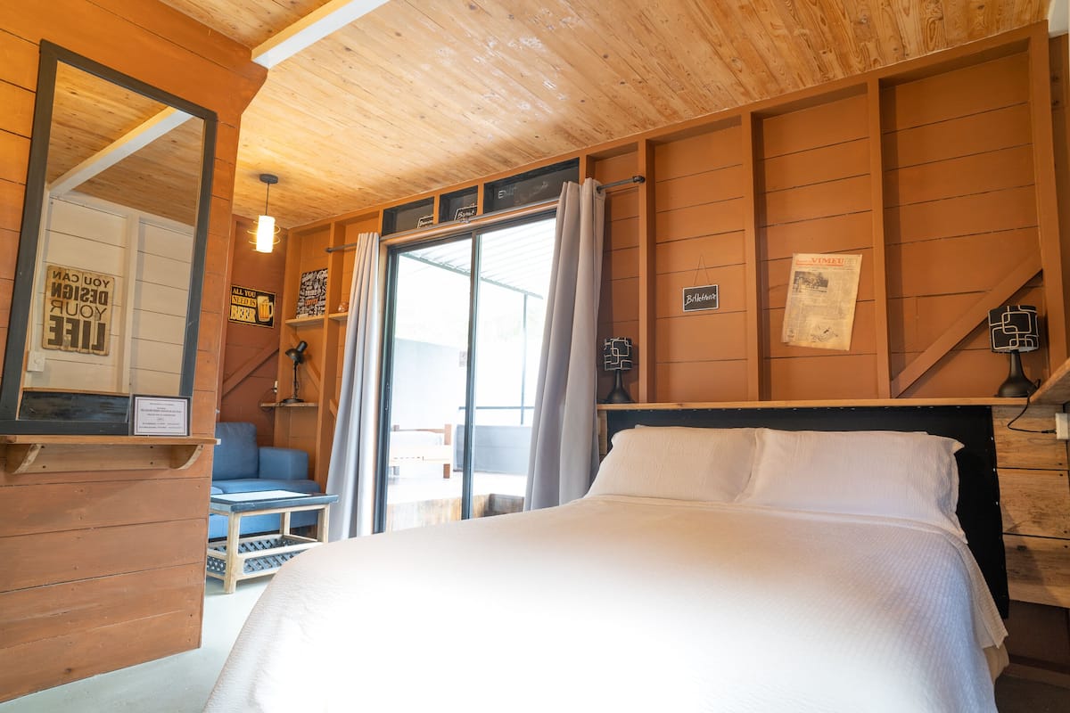 A bedroom with a bed, a mirror, a window with curtains, a blue chair, and a wooden wall.
