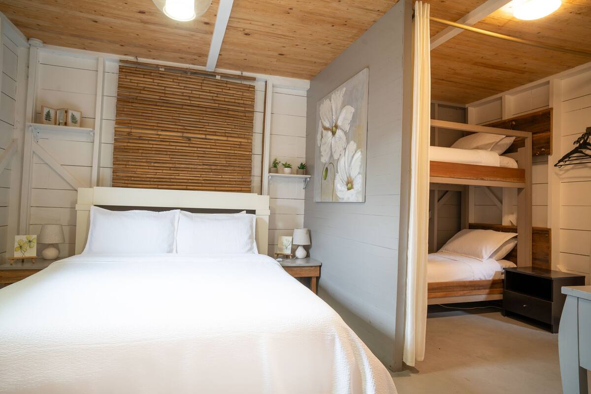 A bedroom with a bed, a nightstand, a lamp, a wooden ceiling, and a wall-mounted shelf.