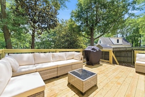 Eine Holzterrasse mit einem Korbsessel und einem überdachten Grill, umgeben von Bäumen.