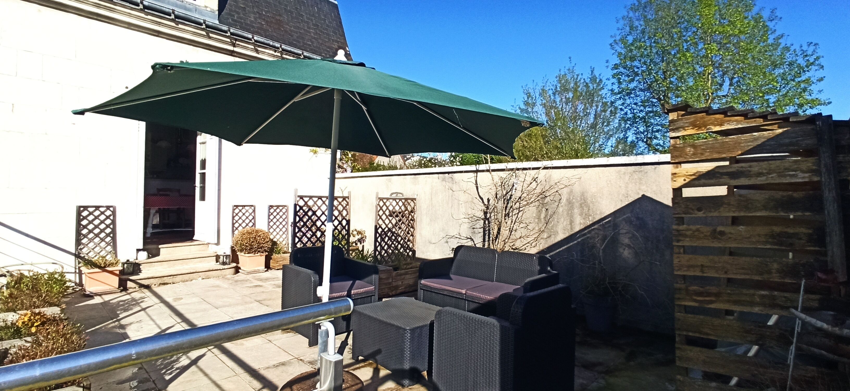 Un espace extérieur aménagé en terrasse, avec un parasol vert, des meubles en osier et une clôture en bois.