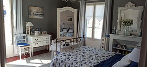 Une chambre à coucher comprenant un lit, une commode, une chaise, une fenêtre avec des rideaux, un miroir et une bibliothèque.