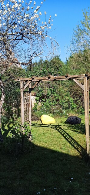 Une pergola en bois dans un jardin, avec un objet jaune et un objet noir sur l’herbe.