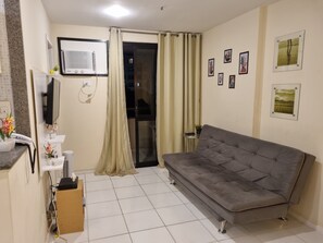 Ein Wohnzimmer mit grauem Sofa, einem an der Wand montierten Fernseher, einem kleinen Tisch mit einer Pflanze und einer Glastür.