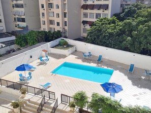 Ein Pool auf dem Dach mit blauen Liegestühlen und Sonnenschirmen, umgeben von Grünpflanzen und Wohnhäusern.