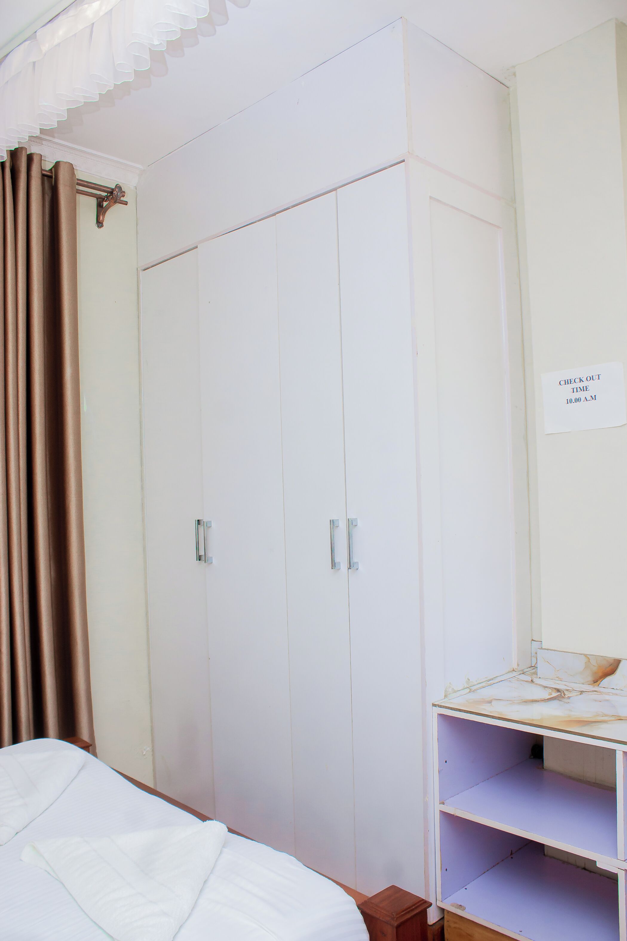 A bedroom with a bed, a wardrobe, and a shelf with a sign indicating check-out time.