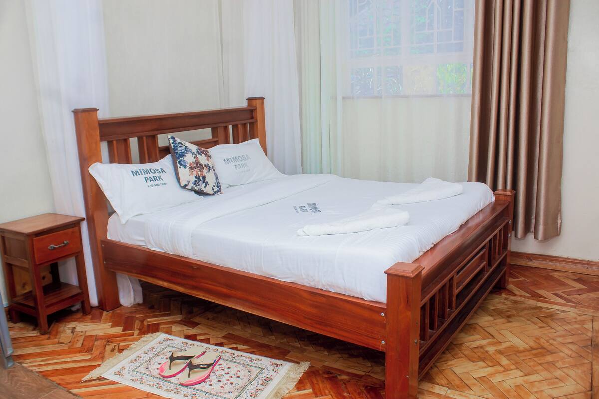 Una cama de madera con ropa de cama blanca, una mesita de noche y un tapete estampado sobre un piso de parquet de madera.