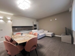 Une cuisine moderne avec des meubles blancs, une table à manger entourée de chaises roses et un canapé d’angle gris.