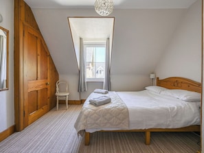 Une chambre avec un plafond en pente, une porte en bois, une chaise, un lit recouvert d’une courtepointe et une fenêtre avec des rideaux.