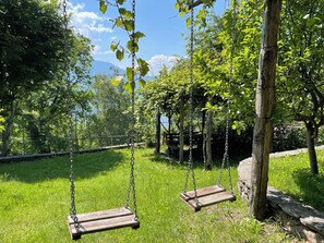 Ein Spielplatz mit einer hölzernen Schaukel, umgeben von Gras, Bäumen und einem Zaun im Hintergrund.