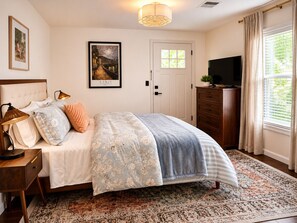Une chambre à coucher avec un lit, des lampes de chevet, un tableau encadré, une porte, une commode, un téléviseur et une fenêtre avec des rideaux.