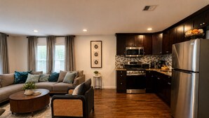 A modern living room with a sofa, coffee table, and a kitchen with dark wooden cabinets, stainless steel appliances, and a tiled backsplash. - Welcome to The Woven Nest Near NYC  (Paterson)