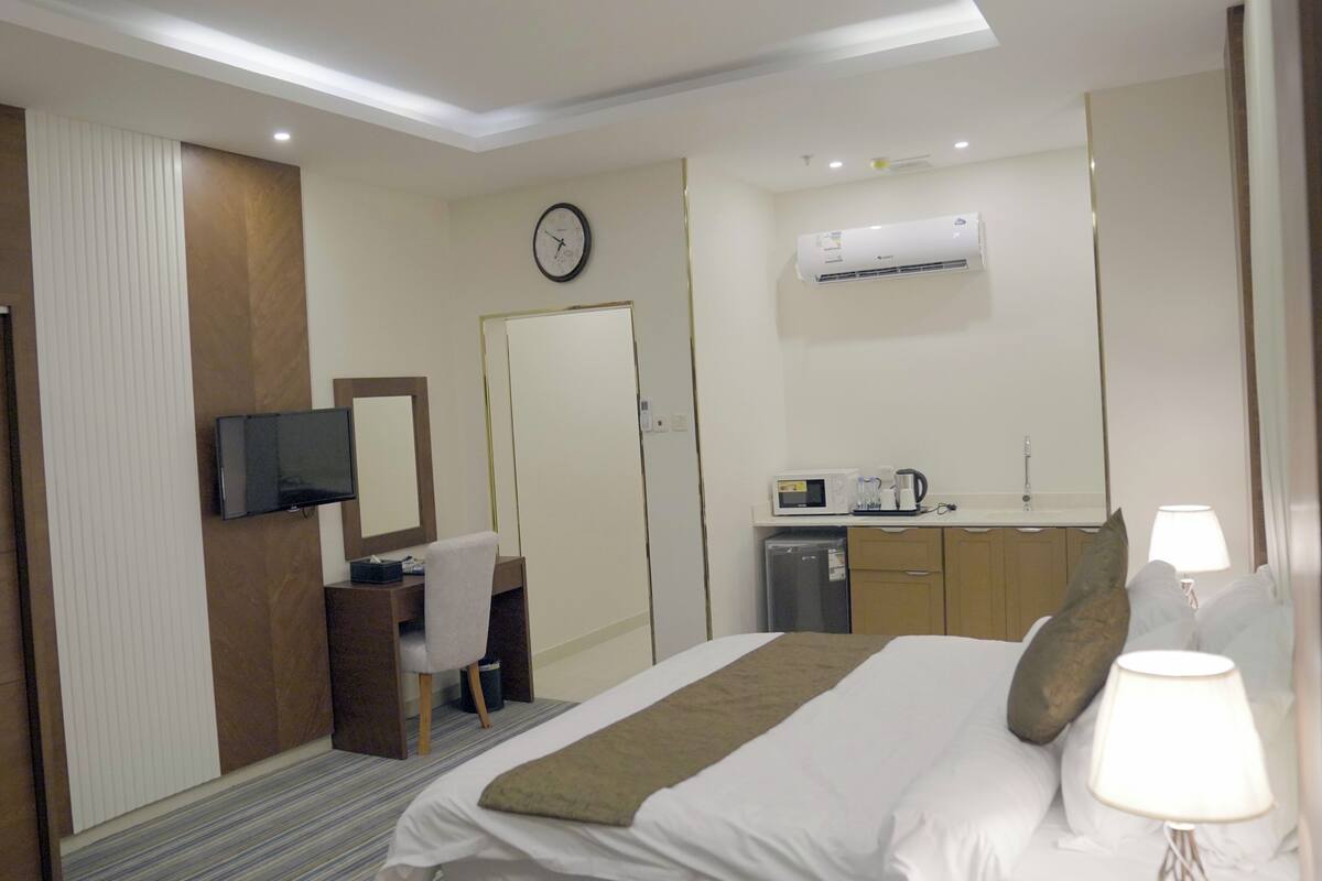 A hotel room with a bed, a desk, a television, a clock, a microwave, and a sink.