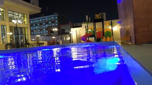 Una piscina con iluminación azul, un tobogán y mobiliario exterior.