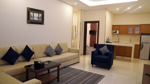 Una sala de estar con sofá, sillón, mesa de centro y una zona de cocina con armarios y electrodomésticos.