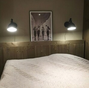 En säng med ett quiltat överdrag, en trägavel och en inramad tavla ovanför. Det finns två vägglampor, en på varje sida om tavlan.