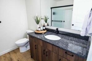 A bathroom with a white toilet, a granite countertop sink, wooden cabinets, and a mirror above the sink. - Welcome to Boise Grove — a thoughtfully designed, modern retreat in one of Boise’s most exciting and fast-growing neighborhoods. Located in Garden City, just steps from greenbelt (Garden City)