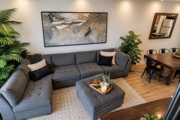 A modern living room with a grey sectional sofa, a dining table, and a large abstract painting on the wall. - Welcome to Boise Grove — a thoughtfully designed, modern retreat in one of Boise’s most exciting and fast-growing neighborhoods. Located in Garden City, just steps from greenbelt, E bikes for rent (Garden City)