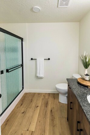 A bathroom with a glass shower door, a toilet, a sink with a granite countertop, and a towel rack. - Welcome to Boise Grove — a thoughtfully designed, modern retreat in one of Boise’s most exciting and fast-growing neighborhoods. Located in Garden City, just steps from greenbelt, E bikes for rent (Garden City)