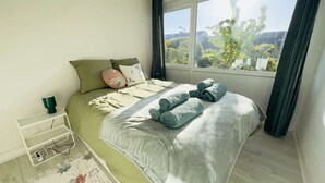 Een slaapkamer met een bed, kussens, een nachtkastje, een raam met uitzicht en een stopcontact.