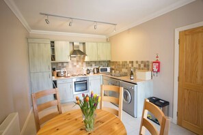 A compact kitchen with modern appliances, a dining table with chairs, and a wooden door. - Holstein Cottage The Dairy at Daviot (Inverness)
