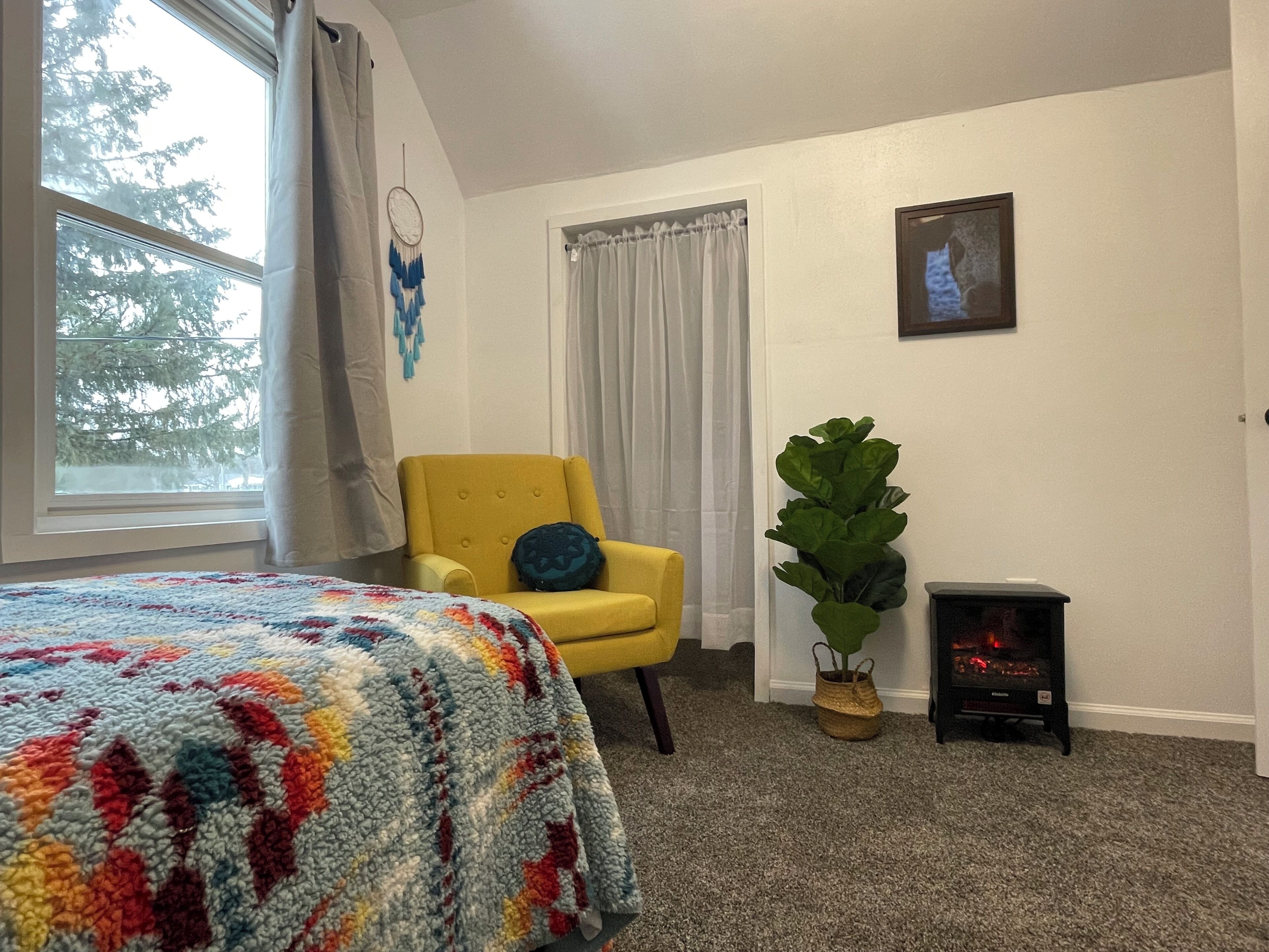 Une chambre à coucher avec un lit, un fauteuil jaune, une grande plante, une cheminée et une fenêtre avec des rideaux.
