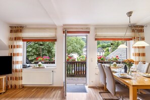 Ein Zimmer mit Esstisch für zwei Personen, einem Fernseher links und Blick auf einen Balkon mit Blumen.