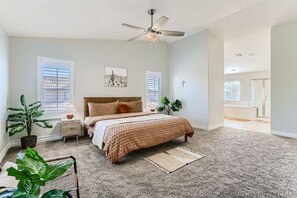 A bedroom with a bed, nightstands, a ceiling fan, a window, and a bathroom visible in the background.