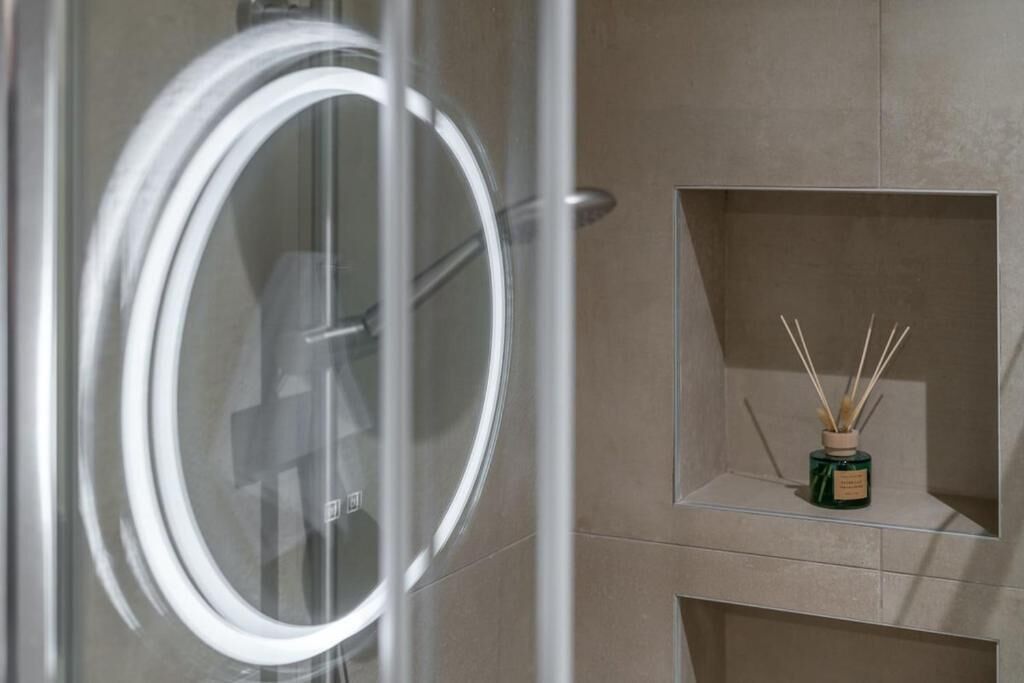 A bathroom with a glass door, a circular mirror, and a shelf with a reed diffuser.