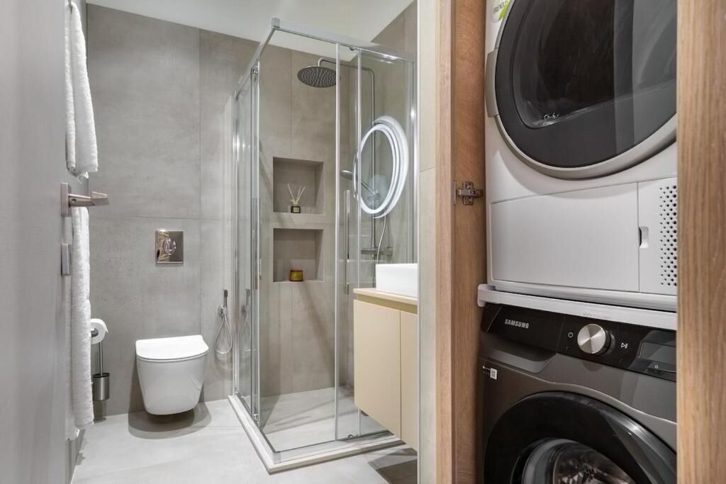 A modern bathroom with a glass-enclosed shower, a toilet, a sink, and a Samsung washing machine.