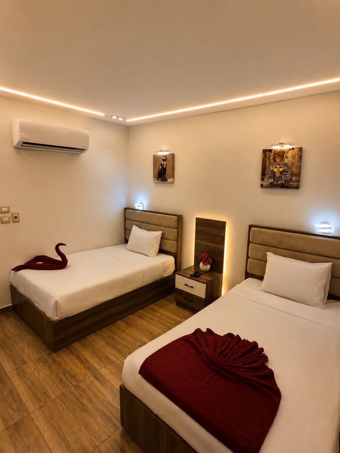 A hotel room with two beds, a nightstand, an air conditioner, and wall-mounted artwork.