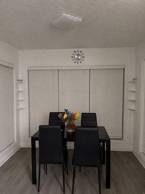 A dining area with a table, chairs, a clock, and a window with blinds.